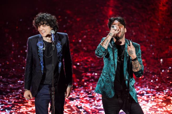 Ermal Meta in piedi in nero vicino a Fabrizio Moro che canta. Alle loro spalle coriandoli rossi