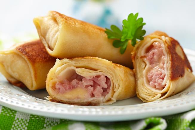 Crepes salate con prosciutto, formaggio e semi di papavero: la ricetta Primo piano si crepes salate con ripieno di prosciutto e formaggio su piatto bianco