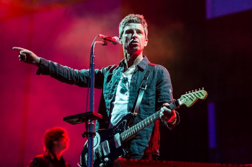 Noel Gallagher agli I-Days Noel Gallagher in piedi, con la chitarra in mano, di fronte a un microfono, con un braccio alzato