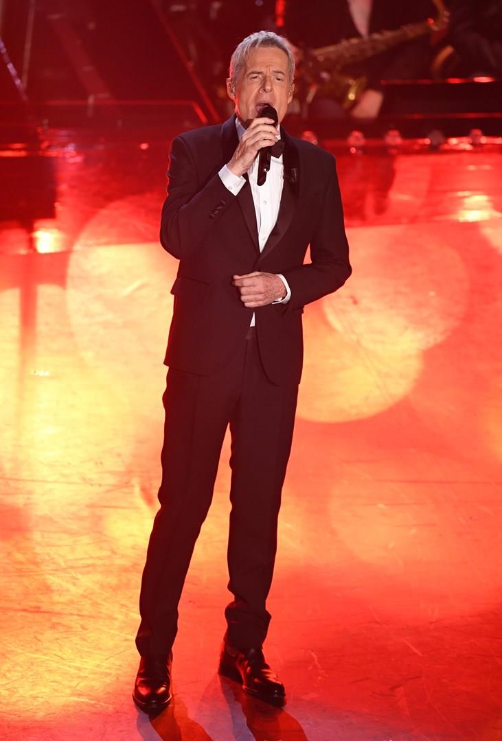 Claudio Baglioni Claudio Baglioni, in piedi, in nero, canta al microfono, su sfondo rosso