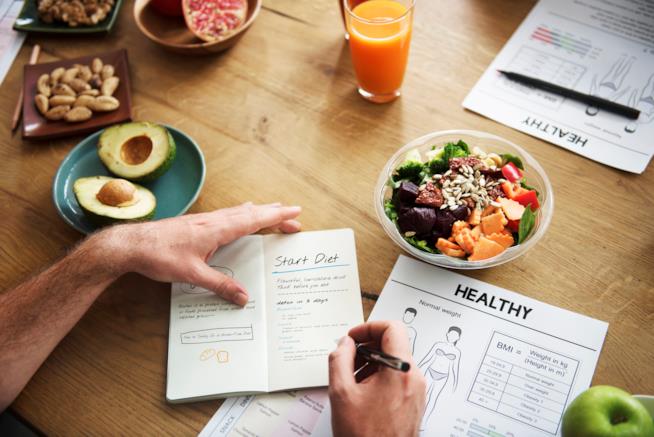 Quali alimenti prevede e cosa promette il regime alimentare TLC studiato in America? Un uomo scrive su un taccuino l'inizio dieta, su un tavolo con frutta, verdura e una spremuta