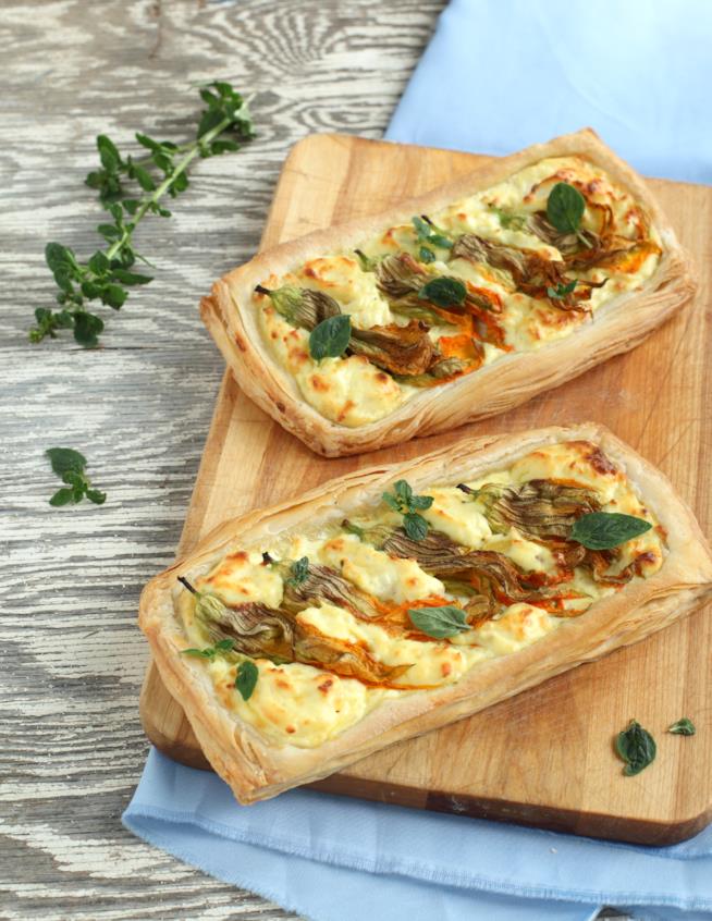Rustico farcito con fiori di zucca e ricotta: la ricetta Tagliere di legno con due rustici salati con ricotta e fiori di zucca, poggiato su tavolo da cucina