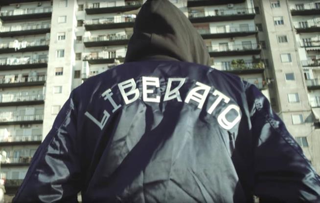 Liberato, il cantante misterioso di cui non si conosce l'identità Liberato, di spalle, con una felpa co la scritta liberato, il cappuccio alzato e lo sfondo di un pal