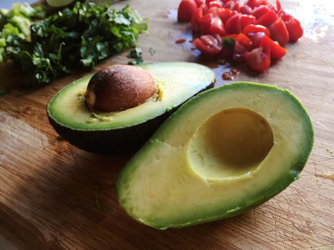 Pregi ed utilizzi dell'avocado, il frutto verde tropicale per eccellenza. un avocado diviso a metà in primo piano, sullo sfondo dei pomodori a fettine e delle erbe