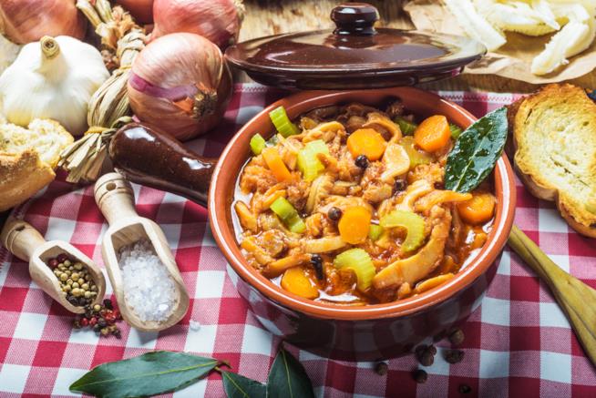 Trippa alla milanese: la ricetta Tegame in terra cotta con all'interno trippa e verdure su tavola a quadri bianca e rossa con ingredi