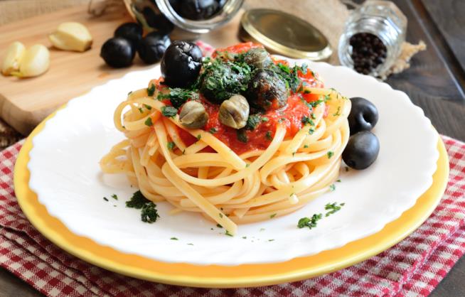 Linguine con capperi e olive: la ricetta Piatto con linguine, capperi, olive nere e prezzemolo in un piatto bianco e giallo e su tovaglia