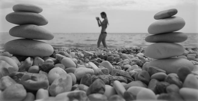 L'estate è in arrivo, ecco i segreti per superare la prova costume Bianco e nero, spiaggia di sassi in primo piano, sullo sfondo una ragazza in bikini in acqua