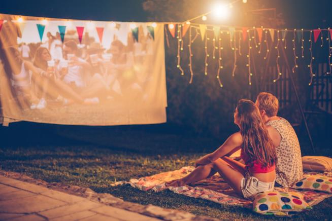 Cinema e primo appuntamento: cinque titoli di film per fare innamorare subito il vostro partner due ragazzi sono seduti all'aperto su una coperta in un prato davanti ad un film proiettato