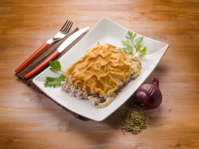 Lasagna bianca con pane carasau: la ricetta Piatto bianco rettangolare con lasagna bianca e qualche foglia di prezzemolo, origano e una cipolla