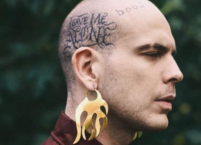 Il rapper Gemitaiz Gemitaiz di profilo, con dei grandi orecchini e i tatuaggi sulla testa, su uno sfondo verde