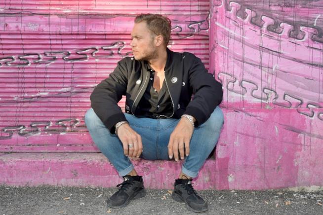 Davide Rossi, figlio di Vasco, debutta nella musica Davide Rossi, giacca e scarpe nere e jeans, seduto, di profilo, davanti a un muro viola