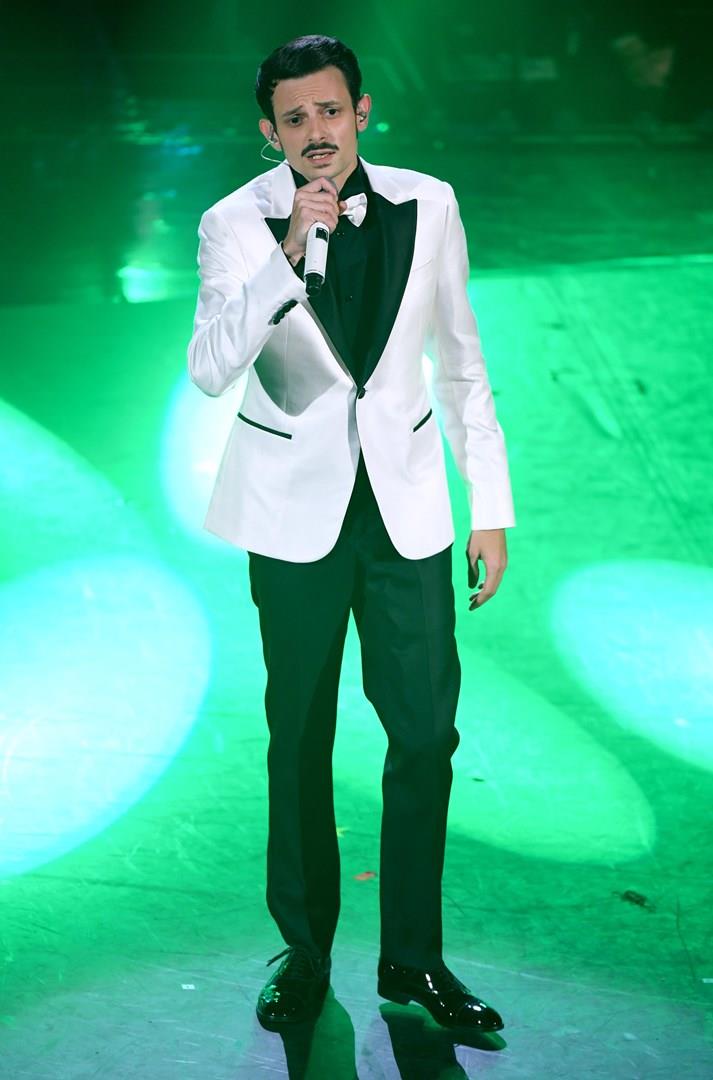 Fabio Rovazzi Fabio Rovazzi, con pantaloni neri e giacca bianca, canta in piedi al microfono su sfondo verde