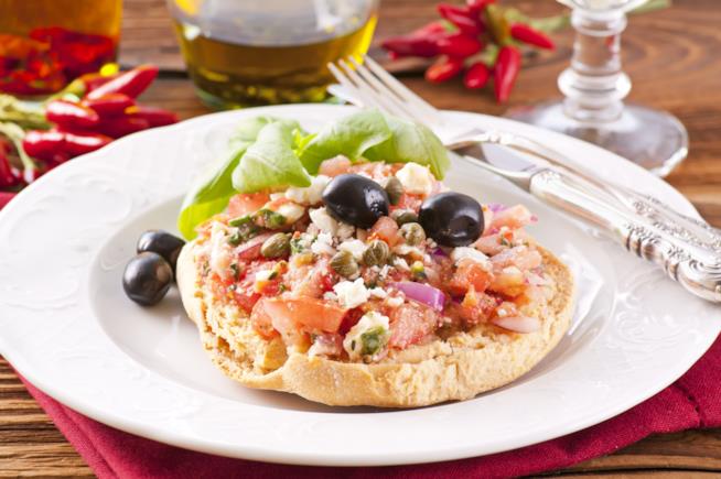 Fresella con tonno, verdure e olive nere: la ricetta Fresella su piatto bianco con pomodori, cipolle, olive nere, tonno, mozzarella e basilico