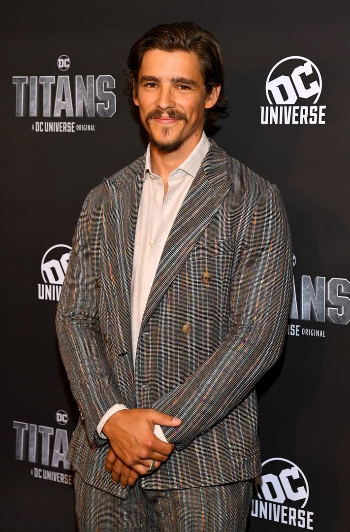 Brenton Thwaites sarà ancora in Titans ma indosserà ancora i panni di Robin? Brenton Thwaitesin giacca e camicia su sfondo pubblicitario di DC Universe e Titans