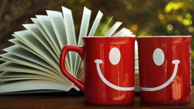 10 libri divertenti da leggere: la classifica Due mug rosse che formano uno smile con un libro aperto da sfondo