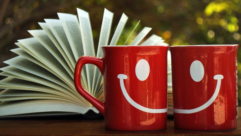 10 libri divertenti da leggere: la classifica Due mug rosse che formano uno smile con un libro aperto da sfondo